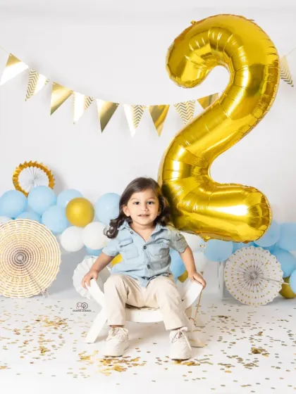 A series of portraits celebrating a second birthday. The toddler poses with a giant gold '2' balloon, surrounded by blue and white balloons and confetti for a festive look.