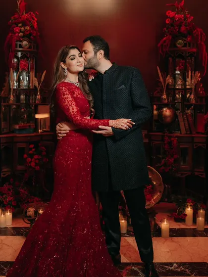 The couple poses in a vintage-themed photo corner, filled with antique props and rich red floral arrangements.
