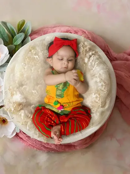 A bright and cheerful photo of a baby girl in a traditional-style outfit. The setup uses a simple bowl prop with flowers to keep the focus on the baby.