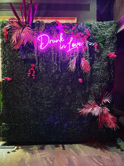 A trendy photo booth for a bachelorette or engagement party. This setup features a lush green wall decorated with pink tropical leaves and a bold "Drunk in Love" neon sign, perfect for stylish party pictures.
