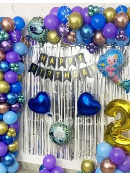 A mermaid-themed 2nd birthday with a silver fringe backdrop. The purple, blue, and gold balloon arch includes a mermaid and heart-shaped foil balloons.