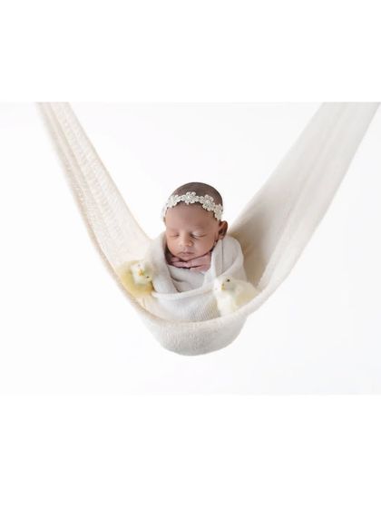 A newborn baby sleeps soundly, swaddled in a soft hanging hammock. The all-white, minimalist setup creates a feeling of floating on a cloud, emphasizing the baby's peaceful and angelic nature.