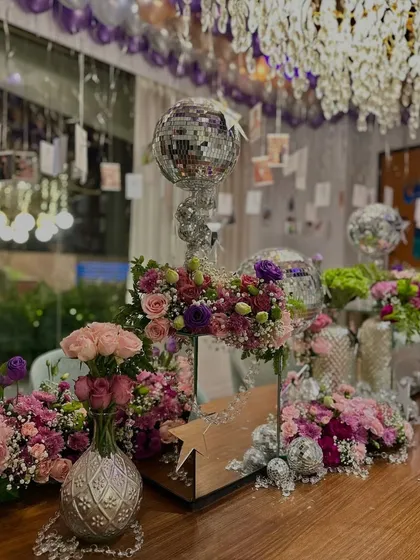 A close-up of the tablescape for a glamorous birthday party. The decor includes intricate floral arrangements in pink and purple, mini disco balls, and silver vases, all reflecting the light from the crystal chandelier above.