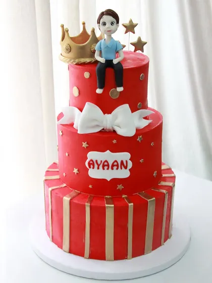 A bold and striking three-tiered red and gold cake for a special boy's birthday. The design features a crown, a big white bow, and a custom name plaque.