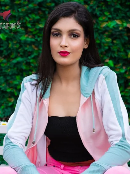A seated outdoor portrait of a female model in a casual, colorful outfit. The green hedge background and soft daylight create a relaxed and appealing image for a fashion portfolio.
