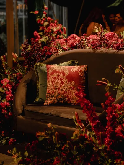 A detail shot of the seating at the red Sangeet. A plush sofa is surrounded by an abundance of red flowers, with a baroque-patterned cushion adding a touch of classic elegance.
