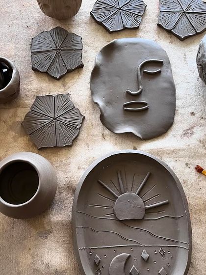 A top-down view of my raw clay creations, including a trinket dish with a sun and moon design, a face plaque, and textured coasters.