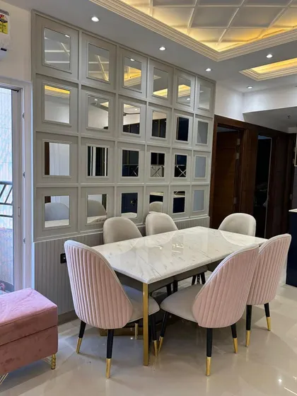 A beautiful dining setup featuring plush pink and beige velvet chairs around a white marble-top table with gold-finished legs. The mirrored grid wall behind the dining area not only adds a decorative element but also reflects light, making the entire space feel larger and brighter.
