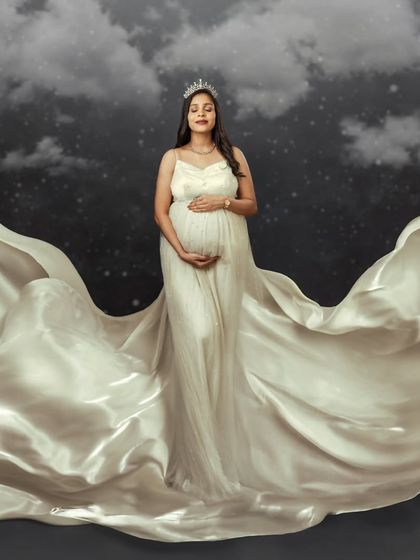A dreamy and artistic portrait of a mom-to-be in a white gown with a massive satin train, set against a cloudy, ethereal backdrop.