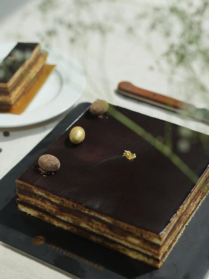 My Opera Cake sits beside a fresh cup of coffee, the perfect pairing. The dark, glossy surface reflects the light, hinting at the complex flavors of coffee and chocolate inside.