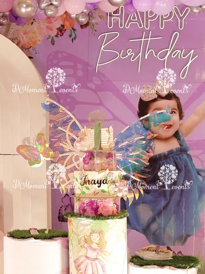 A close-up of the cake table at Inaya's fairy-themed party. The custom-tiered cake is perfectly coordinated with the backdrop, which features a larger-than-life photo of the birthday girl with fairy wings.