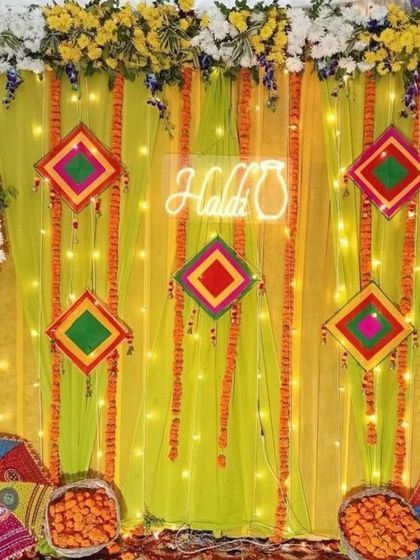 A bright Haldi backdrop with a neon sign and potli. The yellow drape is decorated with colorful geometric hangings and marigold strings, with Rajasthani umbrellas at the base.