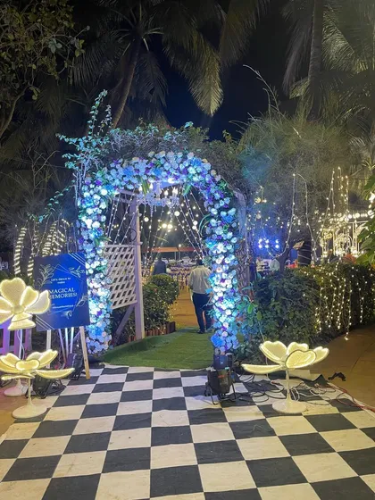 A whimsical entrance for a reception, featuring a checkered path and an arch decorated with blue-lit flowers. The glowing lotus floor lamps add a unique and magical touch.