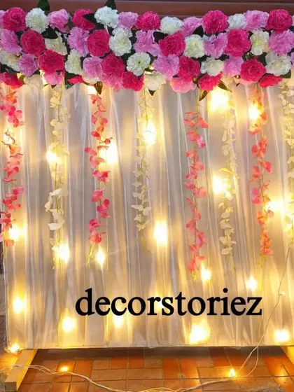This ready-to-install backdrop comes with pre-installed lights that shine through a sheer white curtain. The top is adorned with pink and white carnations, and delicate flower strings hang behind the curtain for a magical effect.