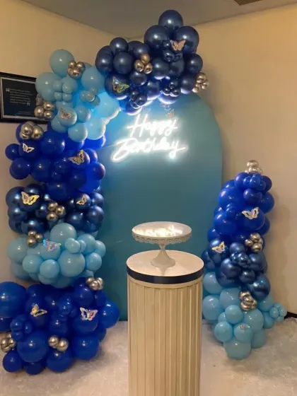 A stylish butterfly-themed birthday setup in shades of blue. The arched backdrop is framed by a beautiful arrangement of blue and silver balloons, with a 'Happy Birthday' neon sign.