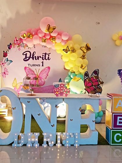 A large-scale butterfly-themed first birthday. The setup includes a 'ONE' marquee table, a photo collage board, and colorful balloon columns.