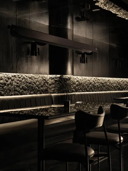 The linear dining arrangement, where a long banquette is set against a textured stone wall. The repetition of tables and chairs creates a strong rhythm, contrasted by the organic nature of the stone.