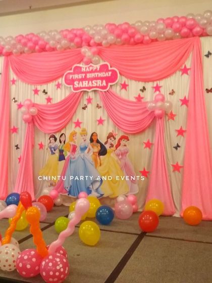 A grand princess themed first birthday stage decoration. The pink and white drapes are accented with a balloon arch and cutouts of multiple Disney princesses.