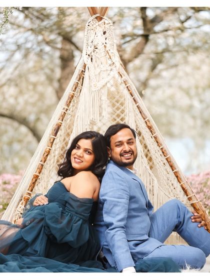 A playful and happy couple portrait, sitting back-to-back against a boho teepee prop outdoors. Their smiles show the joy and excitement of their pregnancy adventure.