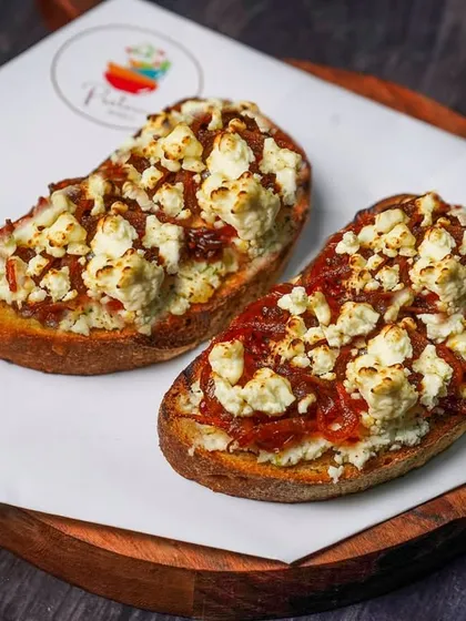 Two pieces of bruschetta with a savory topping of feta cheese and onion jam, served on a wooden board. The dark background creates a cozy, rustic ambiance perfect for an Italian eatery.