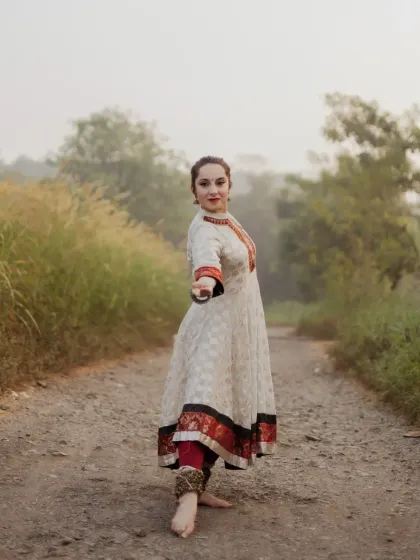 A dynamic shot of the dancer in a powerful Kathak stance. Her direct gaze and outstretched hand invite the viewer into her performance, set against a soft, hazy natural path.