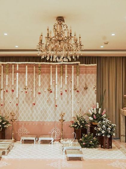 An intimate and traditional engagement setup inside a home. The decor features a backdrop of hanging jasmine garlands and elegant brass lamps, creating a warm and sacred ambiance.