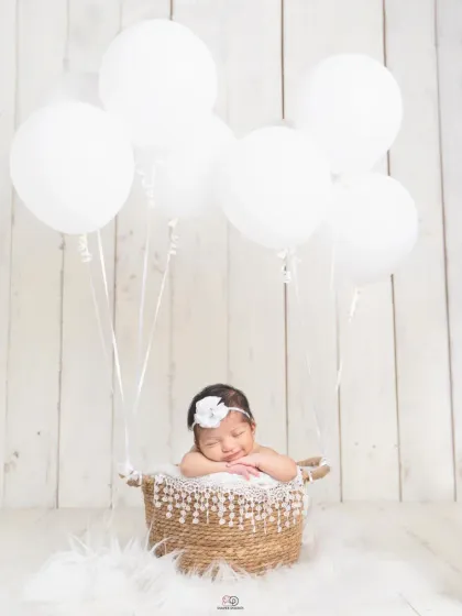 A sleeping newborn rests in a rustic basket, seemingly lifted by a bundle of pure white balloons. The all-white theme creates a clean, minimalist, and heavenly look.