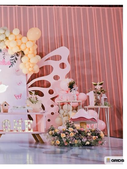 A butterfly-themed first birthday setup in soft pink and peach tones. The decor features a large cutout of butterfly wings, a mushroom house, and a light-up 'ONE' sign.