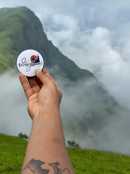 A hand holding our trek badge against the backdrop of Kurinjal peak. These little souvenirs are a reminder of the amazing adventures we have in the Kudremukh range.