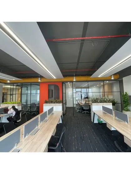 An open-plan workspace with long rows of desks, separated by glass partitions that maintain visual openness. The exposed ceiling with yellow ductwork adds an industrial-chic element to the corporate design.