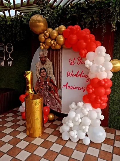 I can personalize your anniversary backdrop with your own photos. This first-anniversary setup features a custom photo print, framed by a red, white, and gold balloon arch, making your memories part of the celebration.