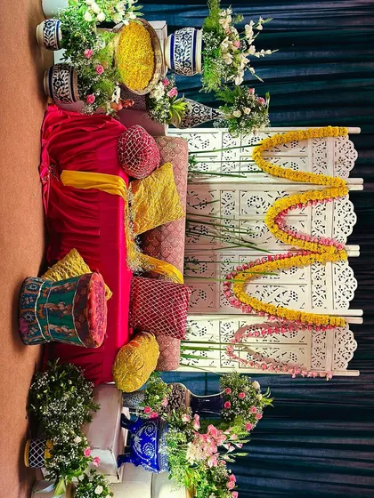 A cozy and colorful seating area for a 'Mehendi Mehfil'. The setup includes a plush sofa with bright cushions, a traditional carved screen, and marigold garlands.