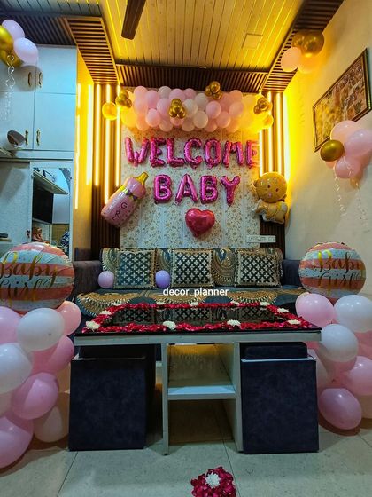 A full room view of a 'Welcome Baby' decoration. The setup includes a balloon backdrop, balloon pillars, and flower petal art on the table, transforming the living room into a party space.
