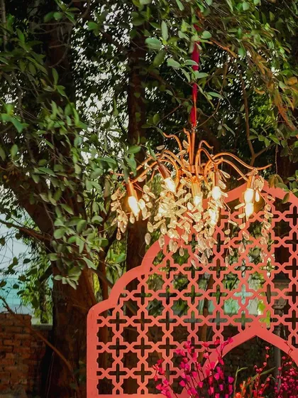 A beautiful shot showing the contrast between the delicate, warm glow of a chandelier and the intricate pattern of a pink jaali screen against the natural backdrop of green trees.