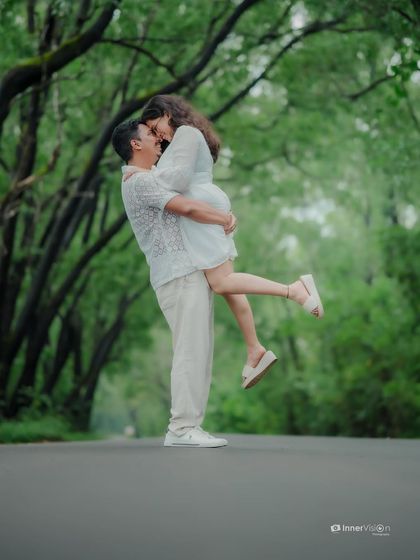 A joyful and spontaneous moment on a road surrounded by a canopy of green trees. The groom lifts his bride-to-be in a playful embrace, capturing pure happiness.