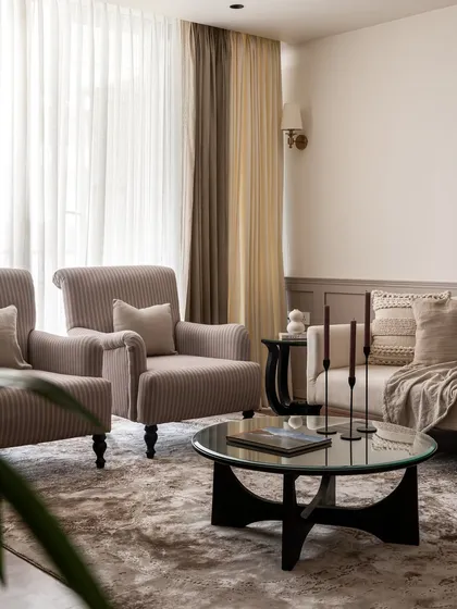The sitting area in Riviera Maison features plush, corduroy-upholstered armchairs and a glass-topped coffee table. The neutral palette and soft textures create a serene and inviting atmosphere for conversation.