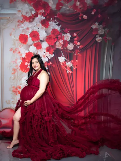 A glamorous maternity portrait of a mother-to-be in a stunning, floor-sweeping red gown against a dramatic floral backdrop.
