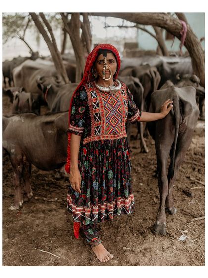 A full-length portrait of a Dhaneta Jat woman in her intricately embroidered dress, standing amongst her herd of buffaloes. This image connects her cultural identity to her daily life.