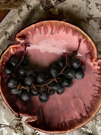 A rich, burgundy-colored ceramic bowl filled with dark grapes, shot from above. The moody, luxurious tones of the bowl and fruit are complemented by the elegant patterned placemat.