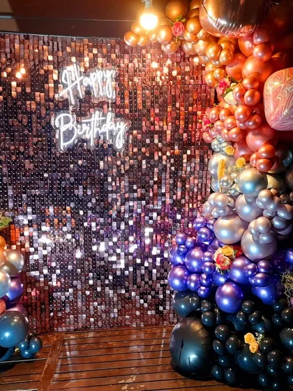 A full view of a dramatic and colorful balloon installation for a birthday party. The organic garland flows from warm oranges and golds to cool silvers, purples, and blacks, creating a striking contrast against the sparkling backdrop.