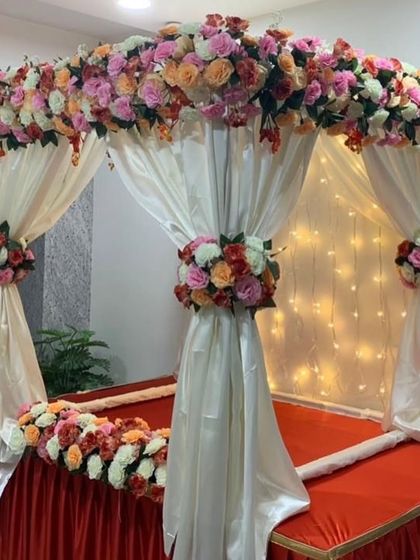 A beautiful four-poster mandap draped in off-white fabric and decorated with bouquets of peach and pink roses. The red base provides a striking contrast.