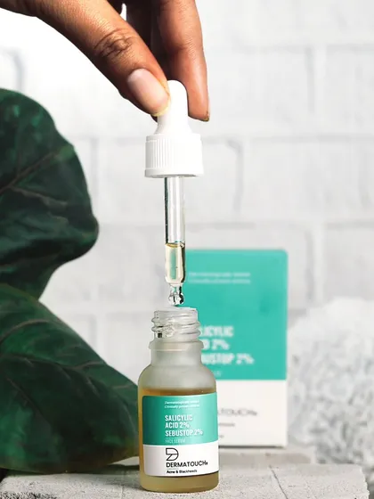 This action shot shows the dropper of a Dermatouch face serum, highlighting the product's liquid consistency and ease of application against a natural, leafy backdrop.