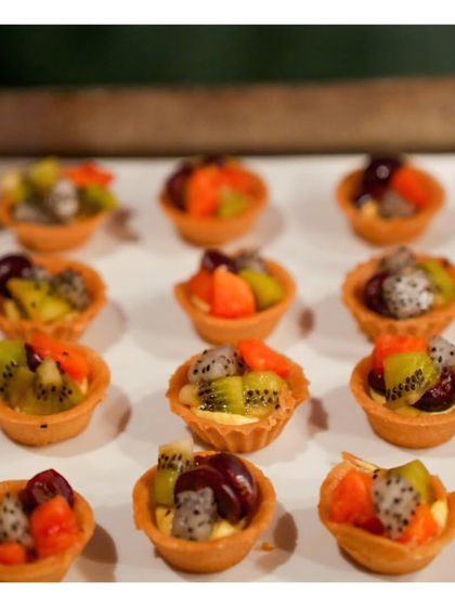 A close up of our miniature fresh fruit tarts. These bite sized pastries feature a crisp shell, creamy custard, and a colourful assortment of seasonal fruits like kiwi, dragon fruit, and grapes.