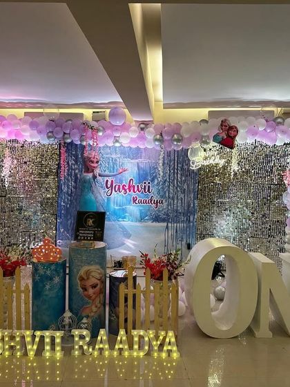 A magical Frozen theme for a first birthday. The stage is decorated with a silver shimmer wall, character plinths featuring Elsa, and a beautiful purple and blue balloon arch.