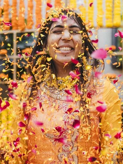 A bride with her eyes closed, smiling in pure bliss as a shower of colorful flower petals rains down on her during her Haldi ceremony. This is a moment of pure, unadulterated joy.