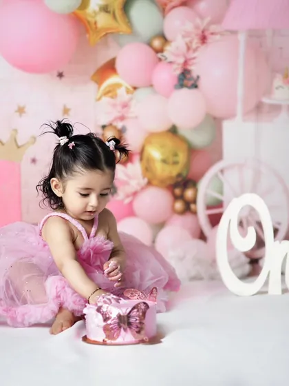 This little princess is delicately touching her butterfly-themed birthday cake. The pink balloon arch and decorations create a dreamy atmosphere for her first birthday celebration.