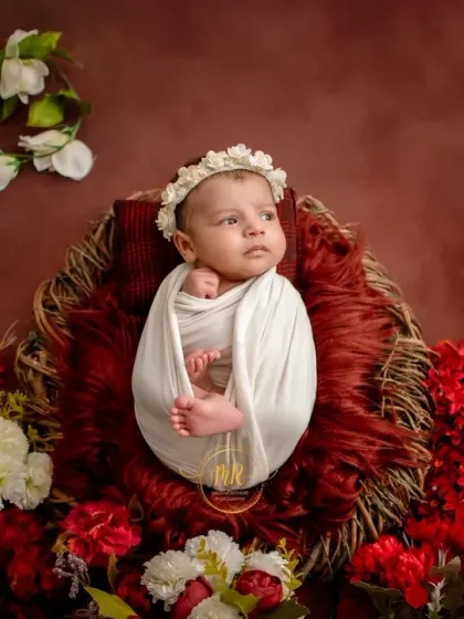 A newborn baby swaddled in white, looking off to the side against a backdrop of vibrant red flowers.