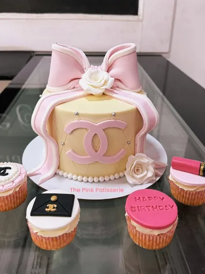 An elegant Chanel-themed cake with matching cupcakes. The main cake features a large pink bow and the iconic logo, while the cupcakes have various brand-related toppers.
