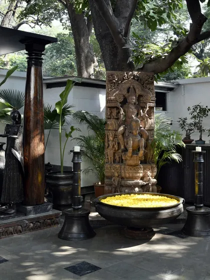 A corner of the courtyard featuring a large stone sculpture of Vishnu and a traditional bronze 'Uruli' filled with flowers. This space is a "museum without walls," where art and artifacts are displayed in an open, natural setting.