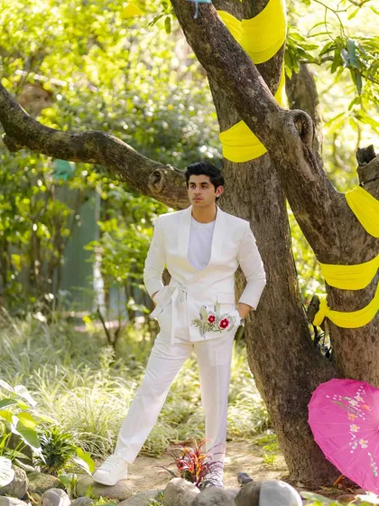 A stylish portrait of the groom-to-be. His all-white ensemble with floral embroidery detail is modern yet traditional, perfectly complementing the colorful, festive decor in the background.